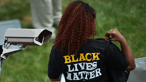 A woman stands with her back to the camera wearing a 'Black Lives Matter' T-shirt. Next to her is a surveillance camera.